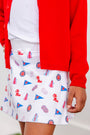 A child wears The Beaufort Bonnet Company’s Ole Miss Searcy Skort – Gawsh Almighty with Worth Avenue White, paired with a white shirt and a bright red cardigan. Only the lower torso and hand are visible against a plain, light-colored background.