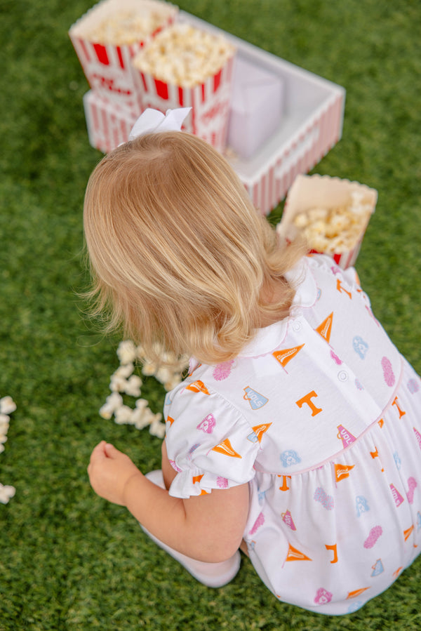 A young child wearing The Beaufort Bonnet Company’s Tennessee Britt Bubble in Victory in Orange with Palm Beach Pink sits on green grass near spilled popcorn and red-striped popcorn boxes.