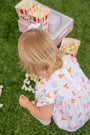 A young child wearing The Beaufort Bonnet Company’s Tennessee Britt Bubble in Victory in Orange with Palm Beach Pink sits on green grass near spilled popcorn and red-striped popcorn boxes.