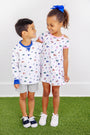 Two smiling children stand on green grass. The boy wears The Beaufort Bonnet Company’s Florida Cassidy Comfy Crewneck – Florida Forever with Florida Blue, paired with gray shorts. The girl, sporting a blue bow, is dressed in a white dress adorned with colorful patterns. They look at each other happily.