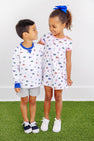 Two smiling children stand on green grass. The boy wears The Beaufort Bonnet Company’s Florida Cassidy Comfy Crewneck – Florida Forever with Florida Blue, paired with gray shorts. The girl, sporting a blue bow, is dressed in a white dress adorned with colorful patterns. They look at each other happily.