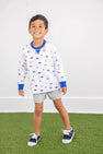 A young boy smiles while wearing The Beaufort Bonnet Company’s Florida Cassidy Comfy Crewneck – Florida Forever with Florida Blue, featuring colorful flag patterns, paired with gray shorts and navy sneakers, standing on green artificial grass against a plain white wall.