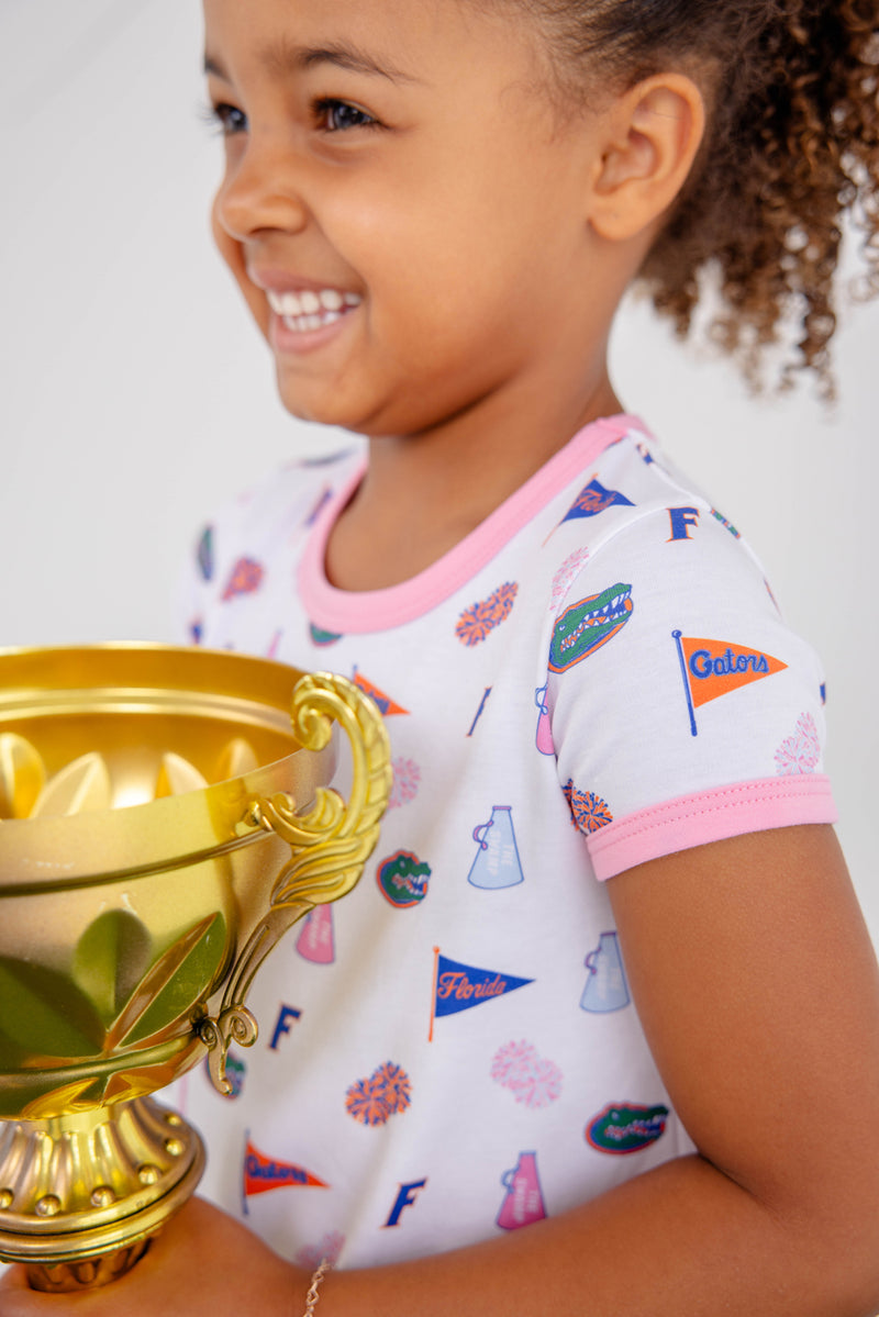 A smiling young girl in The Beaufort Bonnet Company’s Florida Polly Play Dress – Florida Forever with Hamptons Hot Pink print holds a large gold trophy.