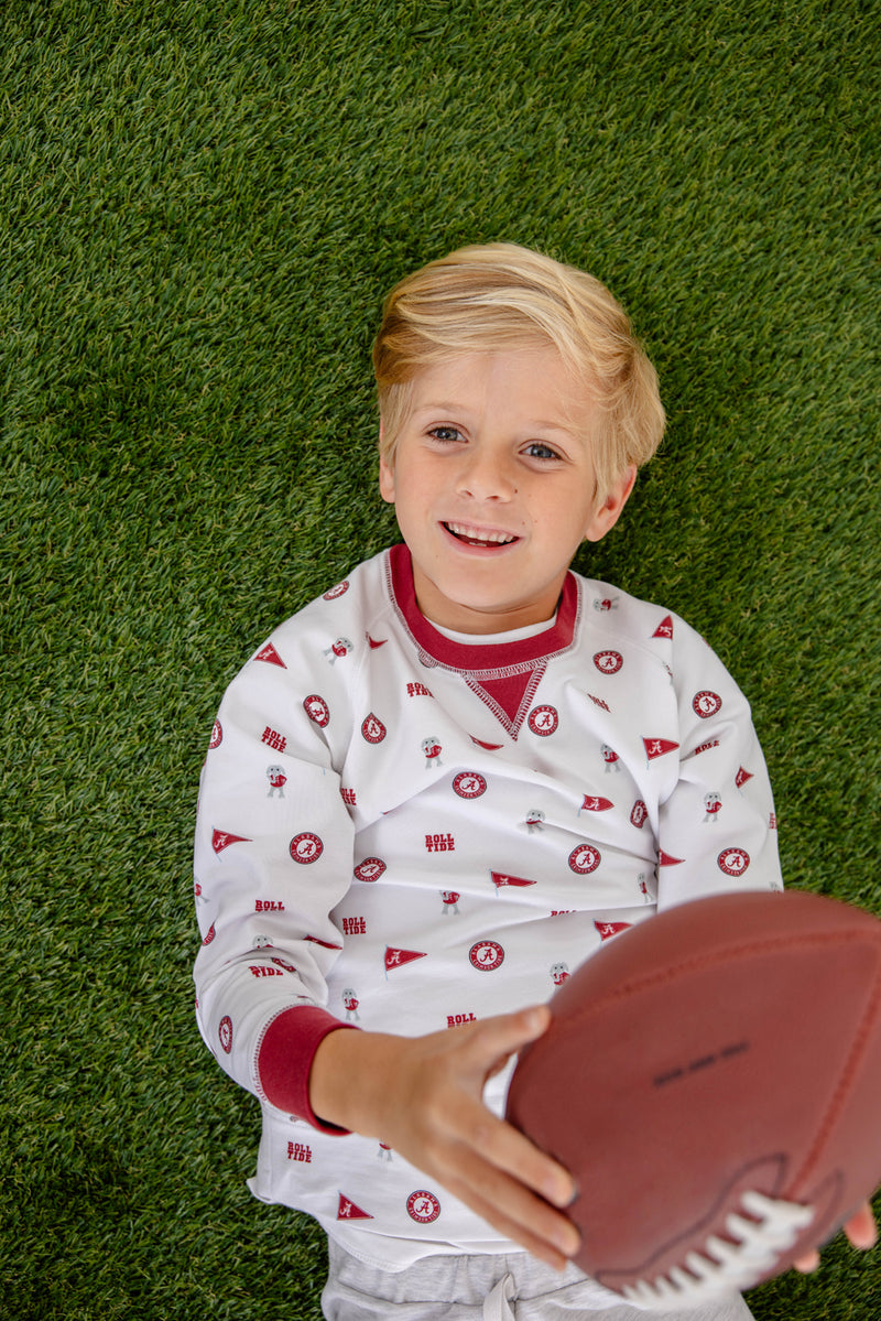 A young boy with blonde hair lies on green grass, smiling and holding a football while wearing The Beaufort Bonnet Company’s Alabama Cassidy Comfy Crewneck in Sweet Home Honey with Alabama Crimson.