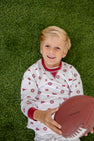 A young boy with blonde hair lies on green grass, smiling and holding a football while wearing The Beaufort Bonnet Company’s Alabama Cassidy Comfy Crewneck in Sweet Home Honey with Alabama Crimson.