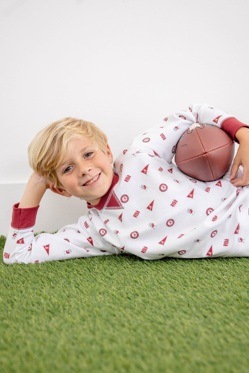 A young child with blonde hair lies smiling on artificial grass, wearing white pajamas with red patterns and The Beaufort Bonnet Company’s Alabama Cassidy Comfy Crewneck - Sweet Home Honey with Alabama Crimson, while holding a small football under their arm.