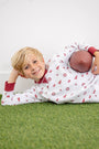 A young child with blonde hair lies smiling on artificial grass, wearing white pajamas with red patterns and The Beaufort Bonnet Company’s Alabama Cassidy Comfy Crewneck - Sweet Home Honey with Alabama Crimson, while holding a small football under their arm.