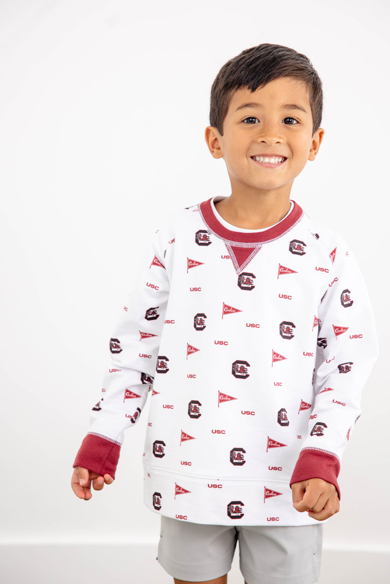 A smiling young boy stands in front of a plain white background, showing off his South Carolina Spirit in The Beaufort Bonnet Company’s USC Cassidy Comfy Crewneck—featuring white fabric with South Carolina Burgundy details and University of South Carolina logos and flags.