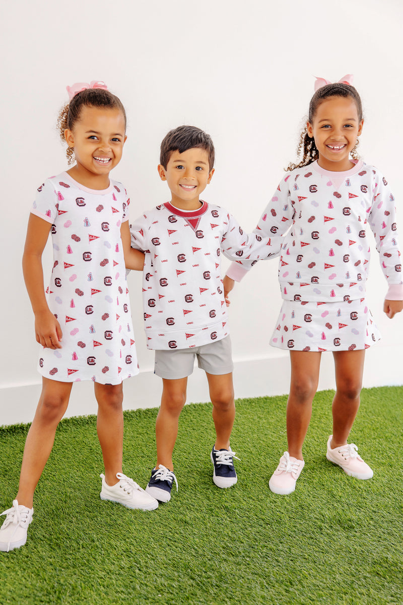 Three smiling young children in white sports-themed outfits stand on green turf against a white wall, holding hands. Their looks feature the South Carolina Spirit: two girls wear hair bows and The Beaufort Bonnet Company’s USC Cassidy Comfy Crewneck – South Carolina Spirit with South Carolina Burgundy—one styled with a dress and the other with a skirt—while the boy wears shorts.