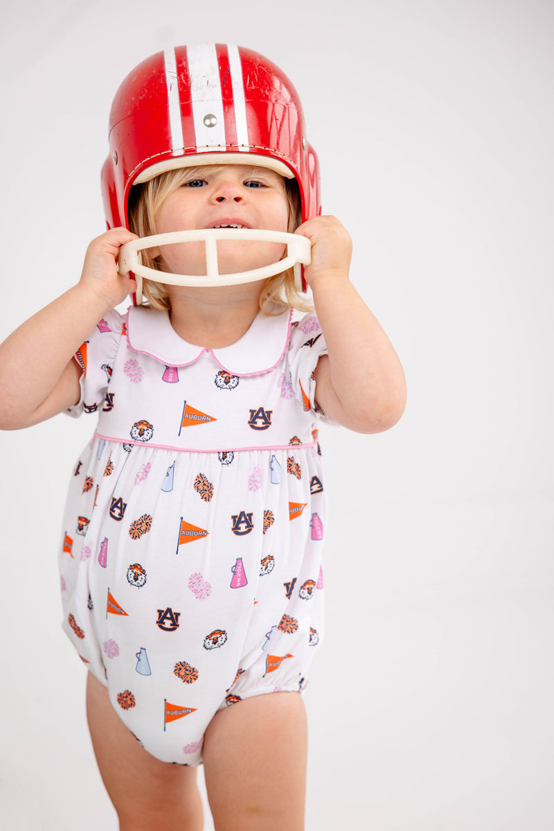 A toddler in a white Auburn Britt Bubble - Tiger Thrill with Hamptons Hot Pink by The Beaufort Bonnet Company, adorned with Auburn University logos and a red football helmet, smiles while holding the helmet’s chin guard.