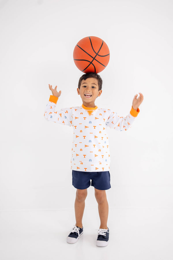Smiling young boy wearing The Beaufort Bonnet Company’s Tennessee Cassidy Comfy Crewneck - Victory in Orange with Tennessee Orange, navy shorts, and sneakers balances a basketball on his head against a plain white background.