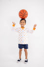 Smiling young boy wearing The Beaufort Bonnet Company’s Tennessee Cassidy Comfy Crewneck - Victory in Orange with Tennessee Orange, navy shorts, and sneakers balances a basketball on his head against a plain white background.