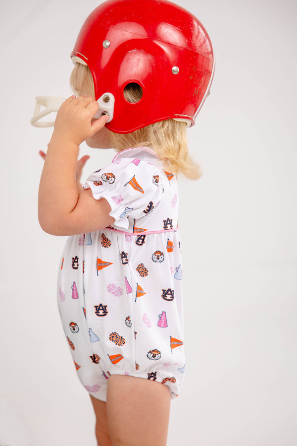 A young child wearing The Beaufort Bonnet Company’s Auburn Britt Bubble - Tiger Thrill with Hamptons Hot Pink, crafted from soft Pima Cotton Knit and featuring Auburn University logos, holds a large red football helmet that partially covers their face while standing against a plain white background.