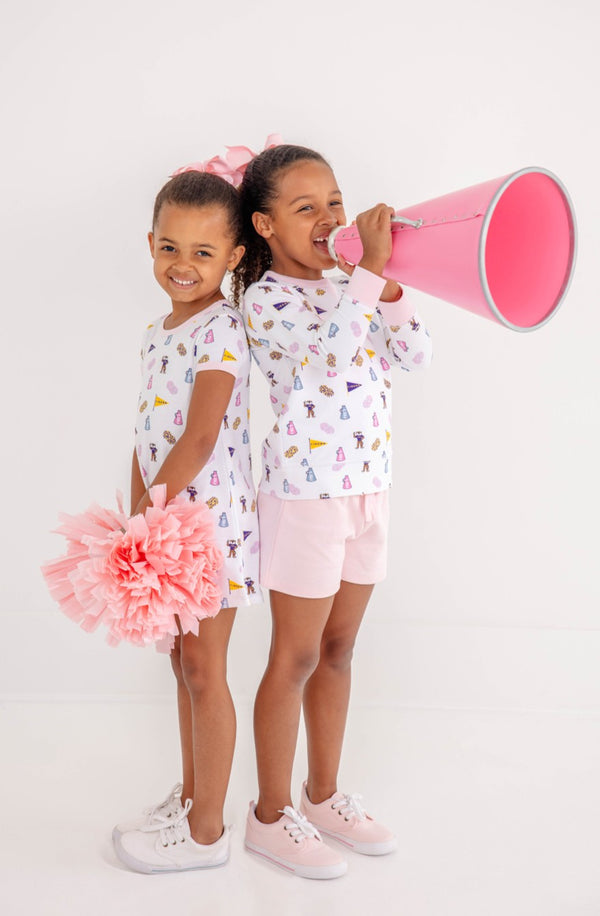 Two young girls stand back to back—one beaming with pink pom-poms, the other cheering through a bold pink megaphone. Both sport matching LSU Polly Play Dress - Tenacious Tigers with Palm Beach Pink by The Beaufort Bonnet Company, styled with classic white shirts, pink shorts, and sneakers, set against a crisp white background for an energetic tribute to Tenacious Tigers spirit.