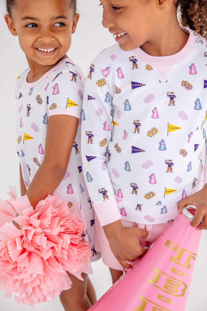 Two young girls smile at each other, wearing matching LSU Polly Play Dresses in the Tenacious Tigers with Palm Beach Pink print by The Beaufort Bonnet Company. One holds pink pom-poms, while the other carries a pink megaphone that says TIBERI.