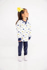 A young girl with a big yellow bow stands smiling and looking to the side against a plain white background, wearing The Beaufort Bonnet Company’s Michigan Cassidy Comfy Crewneck – Bold in Blue with Michigan Blue, paired with navy pants and white sneakers.