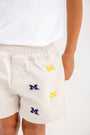 A child is wearing The Beaufort Bonnet Company’s Michigan Critter Sheffield Shorts in Saratoga Stone—a light-colored style featuring embroidered yellow and blue M logos—paired with a white shirt. Only the lower torso and upper legs are visible.