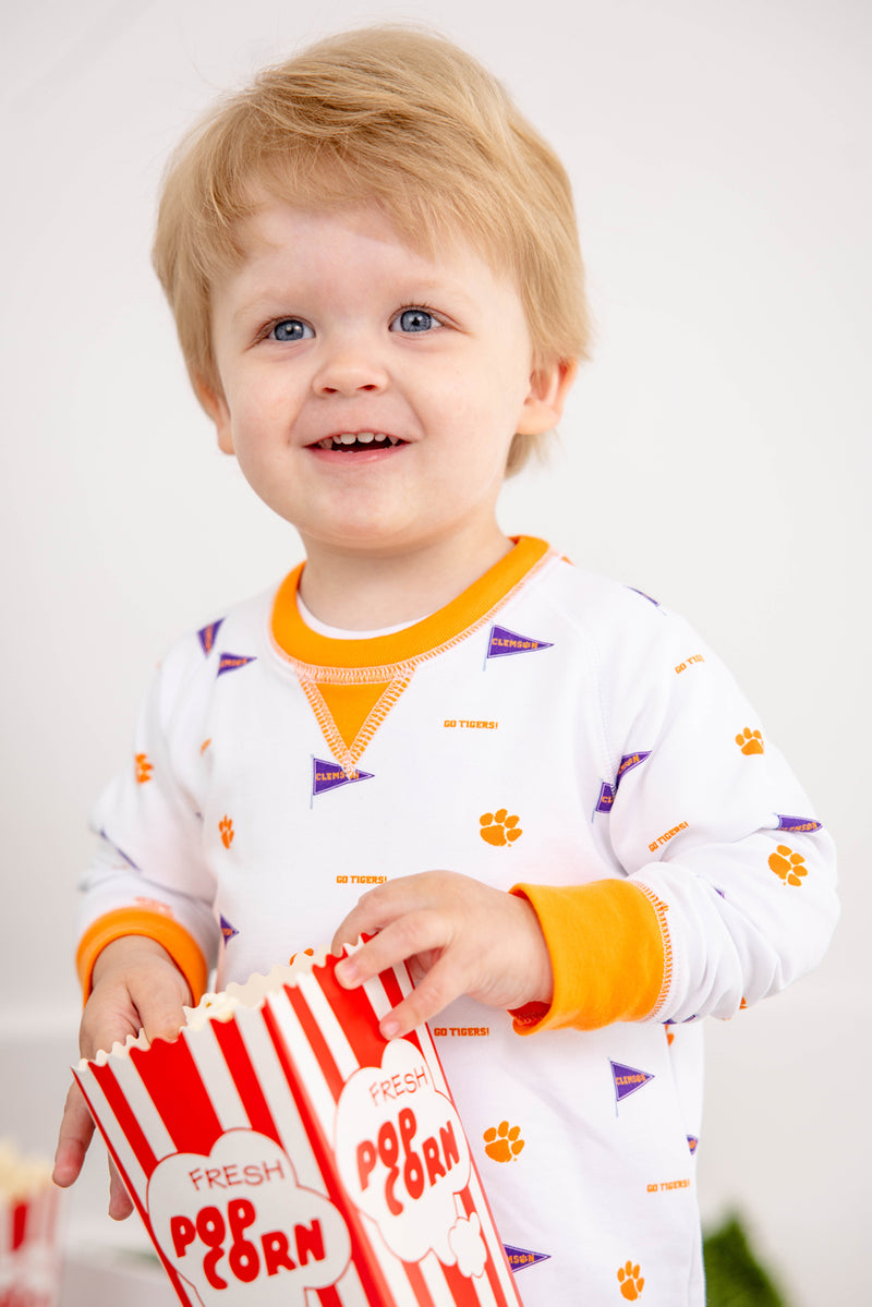A smiling toddler in The Beaufort Bonnet Company’s Clemson Cassidy Comfy Crewneck – Tiger Triumph with Clemson Orange holds a striped carton of popcorn. The bright, simple background draws attention to the child’s joyful expression.