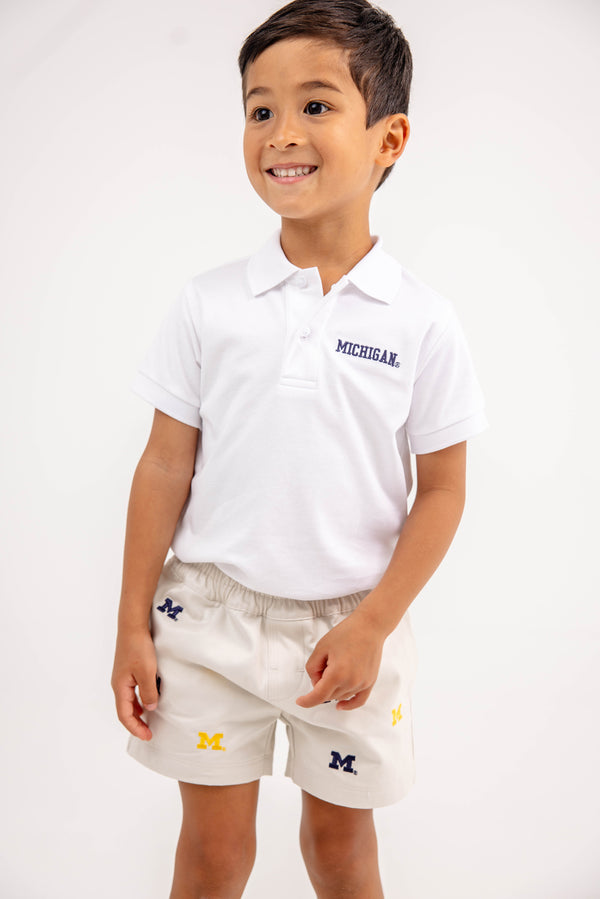 A smiling young boy wears a white Michigan polo shirt and “Michigan Critter Sheffield Shorts - Saratoga Stone” by The Beaufort Bonnet Company, decorated with multiple M logos, standing against a plain white background.
