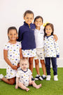 Five children stand and sit together on green turf, smiling brightly. They are dressed in stylish Michigan-themed outfits in blue, yellow, and white, with some wearing The Beaufort Bonnet Company’s Michigan Prim & Proper Polo – Worth Avenue White. Crafted from soft Pima Cotton Knit and adorned with the Michigan logo, these polos add a touch of elegance to their look. One baby sits at the front while the other four children stand behind with arms around each other.