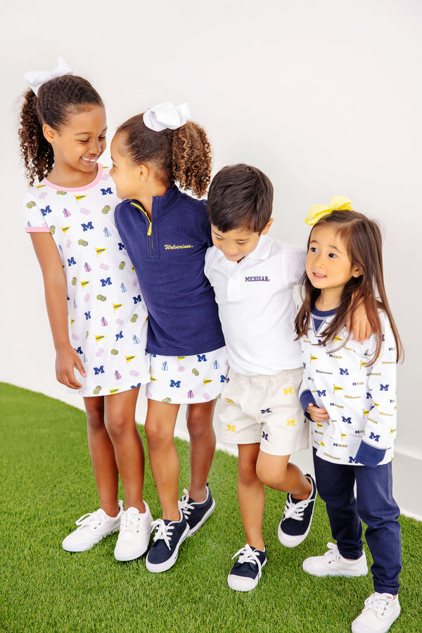 Four young children in Michigan-themed outfits stand together on green turf, smiling and linking arms. Two girls wear white dresses, one girl sports The Beaufort Bonnet Company’s Michigan Hayword Half-Zip in Michigan Blue with Michigan Yellow, and a boy wears a white polo and shorts.