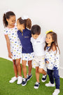 Four young children in Michigan-themed outfits stand together on green turf, smiling and linking arms. Two girls wear white dresses, one girl sports The Beaufort Bonnet Company’s Michigan Hayword Half-Zip in Michigan Blue with Michigan Yellow, and a boy wears a white polo and shorts.