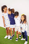 Four young children in Michigan-themed outfits stand together on green turf, smiling and linking arms. Two girls wear white dresses, one girl sports The Beaufort Bonnet Company’s Michigan Hayword Half-Zip in Michigan Blue with Michigan Yellow, and a boy wears a white polo and shorts.