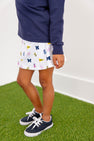 A child wearing navy sneakers, a navy long-sleeve shirt, and The Beaufort Bonnet Company’s Michigan Searcy Skort - Bold in Blue with Worth Avenue White, featuring University of Michigan-themed prints, stands on green artificial grass.