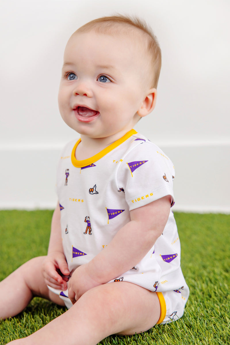 A smiling baby with blue eyes sits on green grass, wearing The Beaufort Bonnet Company’s LSU Short Sleeve Patton Play Bubble - Tenacious Tigers with LSU Gold. The background is plain white.