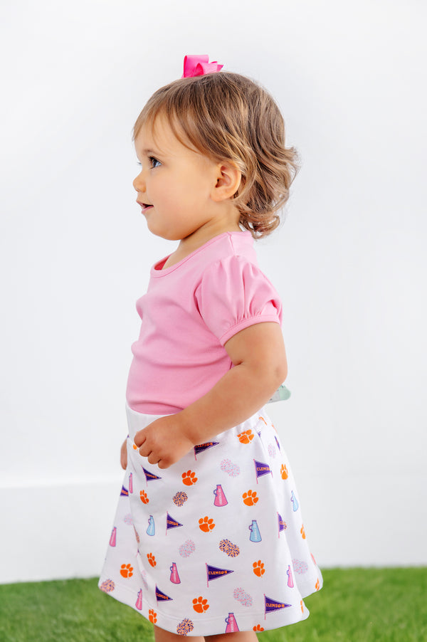 A young child with light brown hair, dressed in a pink shirt and The Beaufort Bonnet Company’s Clemson Searcy Skort - Tiger Triumph with Worth Avenue White, stands on green grass against a plain white background.