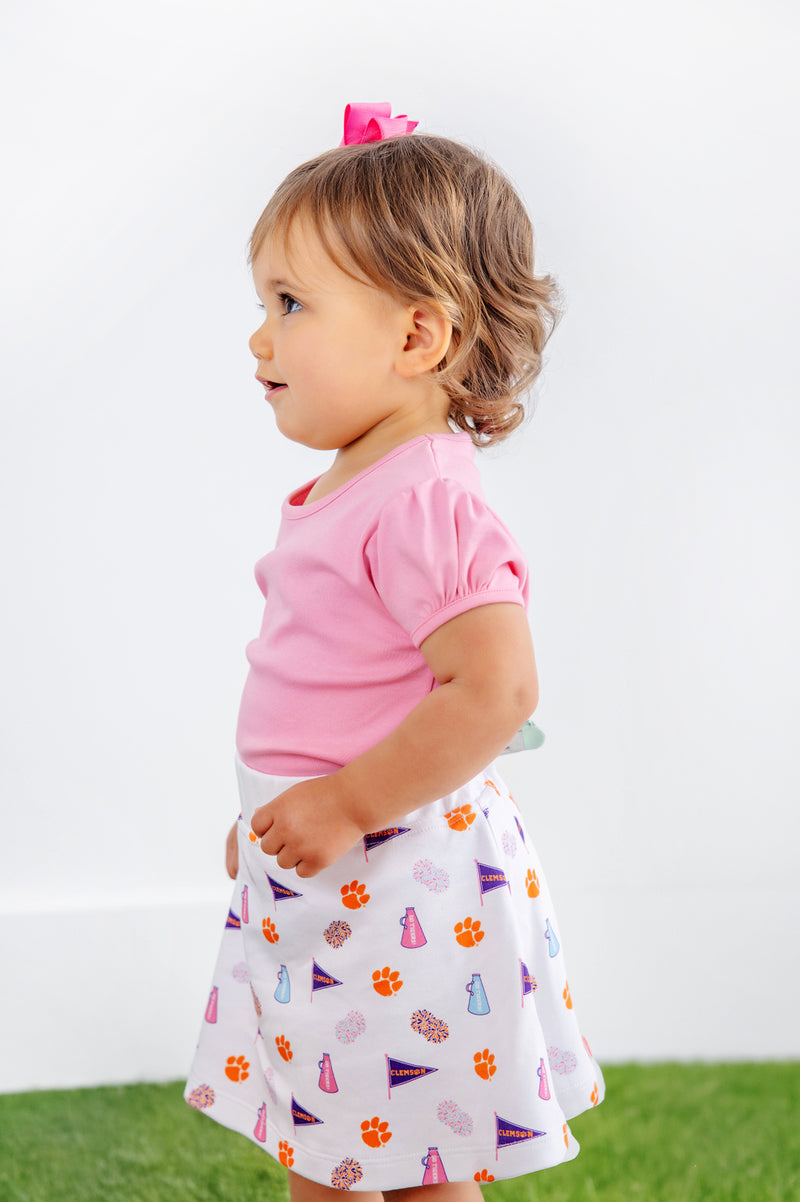 A young child with light brown hair, dressed in a pink shirt and The Beaufort Bonnet Company’s Clemson Searcy Skort - Tiger Triumph with Worth Avenue White, stands on green grass against a plain white background.