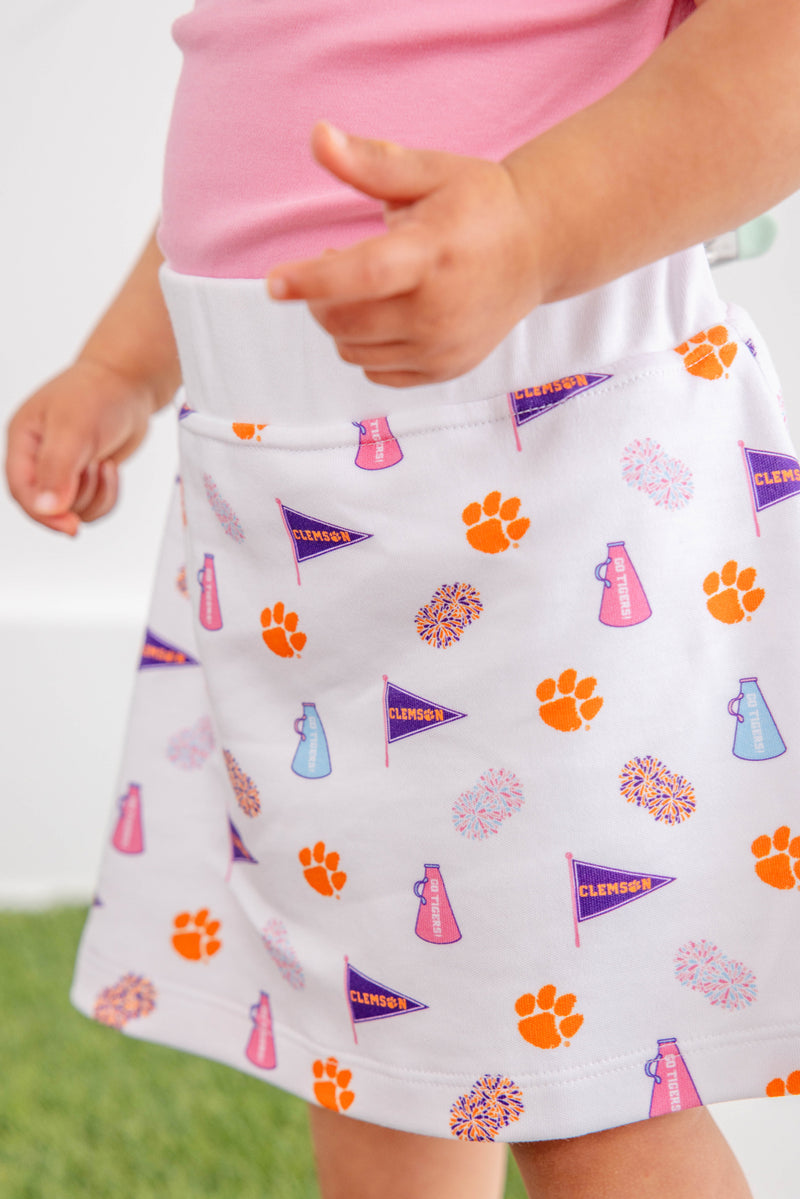 A child is wearing The Beaufort Bonnet Company’s Clemson Searcy Skort – Tiger Triumph with Worth Avenue White, featuring orange paw prints, purple pennants, pink megaphones, and colorful pom-poms. The skort is paired with a light pink shirt, and the background is blurred.