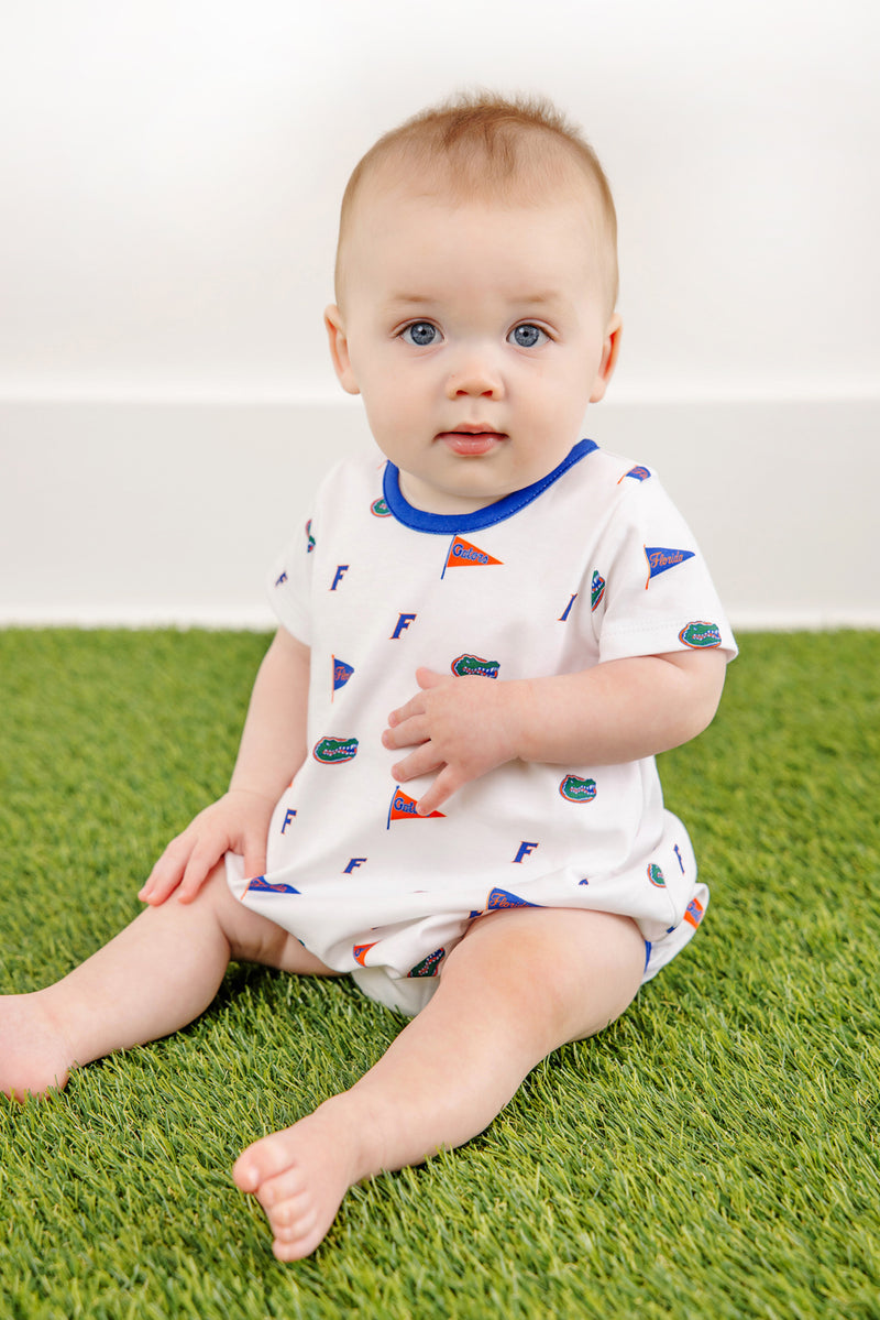 A baby with short light hair and blue eyes sits on green grass, wearing The Beaufort Bonnet Company’s Florida Short Sleeve Patton Play Bubble in the “Florida Forever with Florida Blue” print—a 100% Pima Cotton white onesie featuring blue, orange, and green flags and “F” patterns. The background is simple and white.