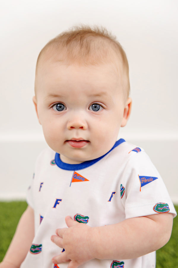 A baby with blue eyes and light brown hair wears The Beaufort Bonnet Company’s Florida Short Sleeve Patton Play Bubble – Florida Forever with Florida Blue, crafted from 100% Pima Cotton and adorned with small Florida Gators logos and flags in the playful Florida Forever print, while sitting against a white background and green floor.