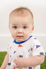 A baby with blue eyes and light brown hair wears The Beaufort Bonnet Company’s Florida Short Sleeve Patton Play Bubble – Florida Forever with Florida Blue, crafted from 100% Pima Cotton and adorned with small Florida Gators logos and flags in the playful Florida Forever print, while sitting against a white background and green floor.