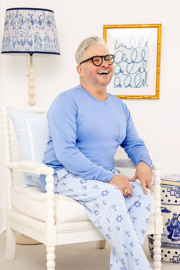 A smiling person with short gray hair and glasses sits on a white chair, wearing The Beaufort Bonnet Company’s Men’s Sunday Style Sweatpants - Eight Nights to Remember, a light blue pajama set adorned with Star of David patterns that radiate holiday comfort. The room is decorated in blue and white accents, including a lamp, pillow, and framed wall art.