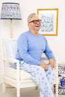 A smiling person with short gray hair and glasses sits on a white chair, wearing The Beaufort Bonnet Company’s Men’s Sunday Style Sweatpants - Eight Nights to Remember, a light blue pajama set adorned with Star of David patterns that radiate holiday comfort. The room is decorated in blue and white accents, including a lamp, pillow, and framed wall art.