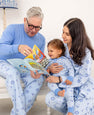 Three people wear The Beaufort Bonnet Company’s “Eight Nights to Remember” Men’s Sunday Style Sweatpants, sitting together and reading a book in matching family outfits. An older man points at the pages while a woman holds a smiling toddler on her lap, all looking engaged and happy in their coordinated holiday loungewear.
