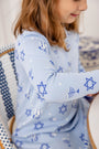 A child wearing The Beaufort Bonnet Company’s “Long Sleeve Polly Play Dress - Eight Nights to Remember,” crafted from soft Pima cotton and adorned with blue Stars of David, menorahs, and dreidels, sits on a woven chair. Smiling and looking down, only the side of her face and upper body are visible in the light blue Hanukkah dress.