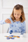 A young girl in The Beaufort Bonnet Company’s Long Sleeve Polly Play Dress – Eight Nights to Remember, adorned with festive Hanukkah symbols, smiles as she spins a dreidel at a white table with chocolate coins, looking down and wearing a white bow in her hair.