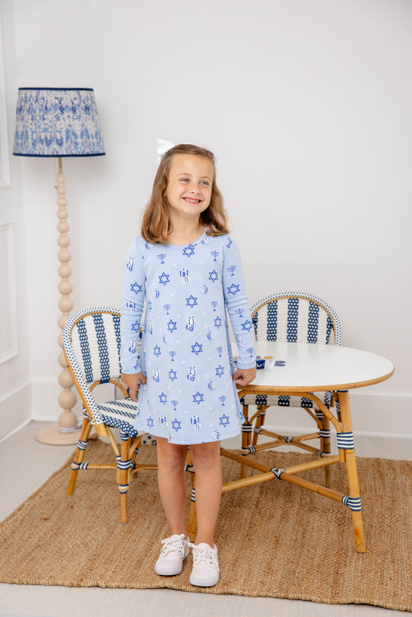 A smiling young girl wears The Beaufort Bonnet Company’s Long Sleeve Polly Play Dress - Eight Nights to Remember, crafted from soft Pima cotton with charming Star of David patterns. She stands in front of a small table and chairs, her look completed by a white hair bow and white sneakers, as a blue-patterned lamp glows in the background.