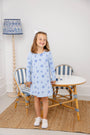 A smiling young girl wears The Beaufort Bonnet Company’s Long Sleeve Polly Play Dress - Eight Nights to Remember, crafted from soft Pima cotton with charming Star of David patterns. She stands in front of a small table and chairs, her look completed by a white hair bow and white sneakers, as a blue-patterned lamp glows in the background.