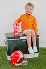 A smiling young boy wears The Beaufort Bonnet Company’s Clemson Hayword Half-Zip in Clemson Orange with Clemson Regalia and Worth Avenue White, sitting on a green cooler while holding popcorn. A red football helmet rests beside him on artificial grass, set against a plain white background.