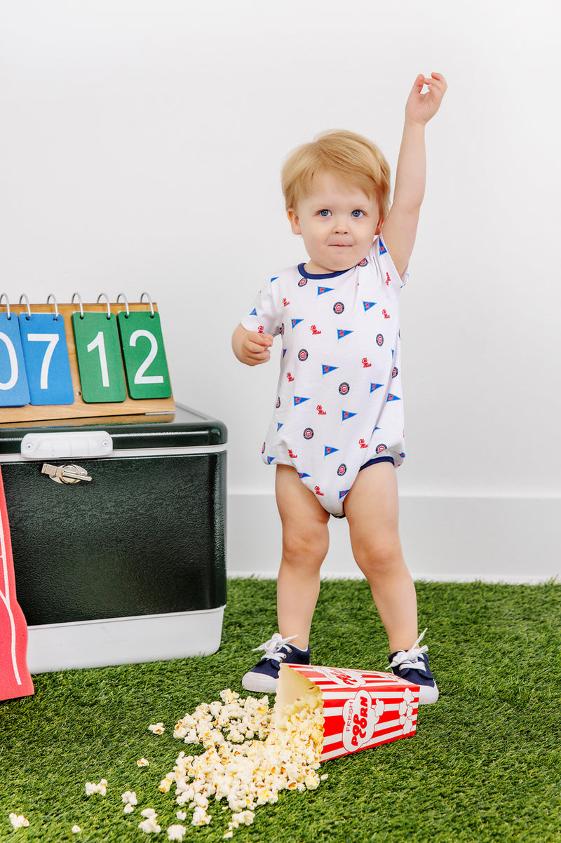 A toddler wearing The Beaufort Bonnet Company’s Ole Miss Short Sleeve Patton Play Bubble - Gawsh Almighty with Nantucket Navy stands on the grass next to spilled popcorn, raising one arm. Behind them are a cooler, number blocks, and a foam finger.