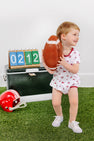 A young child in a white Alabama Short Sleeve Patton Play Bubble - Sweet Home Honey with Alabama Crimson from The Beaufort Bonnet Company holds a toy football while standing on artificial grass. Behind them are a scoreboard displaying 0212, a trunk, and a red football helmet on the ground.