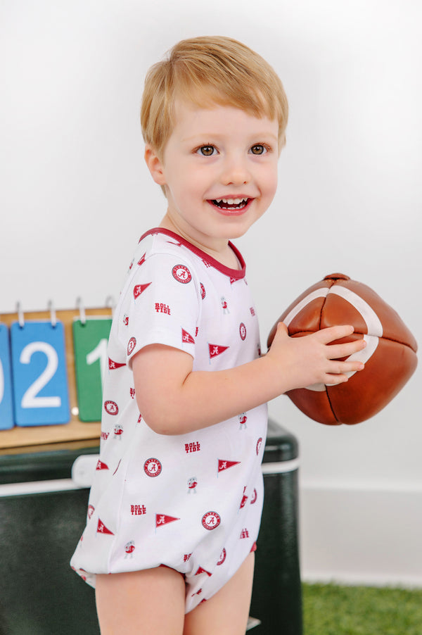 A smiling young child wearing The Beaufort Bonnet Company’s Alabama Short Sleeve Patton Play Bubble in Sweet Home Honey with Alabama Crimson holds a toy football, with a green box and a scoreboard showing the numbers 2, 1, and 0 in the background.
