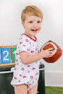 A smiling young child wearing The Beaufort Bonnet Company’s Alabama Short Sleeve Patton Play Bubble in Sweet Home Honey with Alabama Crimson holds a toy football, with a green box and a scoreboard showing the numbers 2, 1, and 0 in the background.