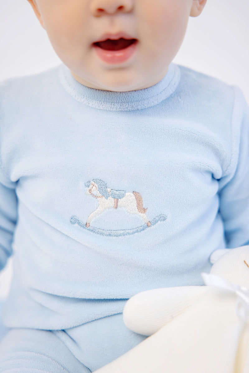 A baby wearing The Beaufort Bonnet Company’s Paxton Playsuit in Buckhead Blue with rocking horse embroidery on the chest holds a white stuffed toy. Only the lower half of the baby’s face is visible, with their mouth slightly open.