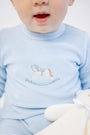 A baby wearing The Beaufort Bonnet Company’s Paxton Playsuit in Buckhead Blue with rocking horse embroidery on the chest holds a white stuffed toy. Only the lower half of the baby’s face is visible, with their mouth slightly open.