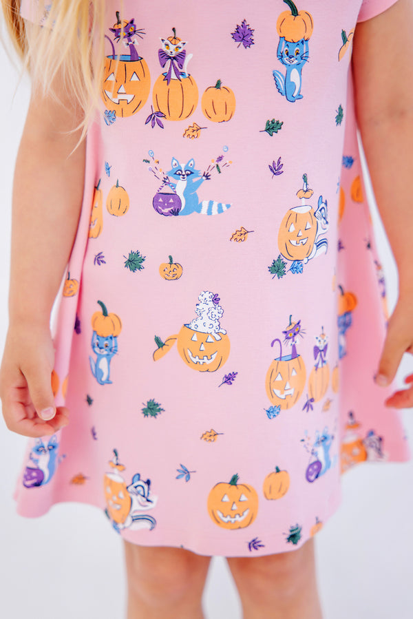 A child wearing The Beaufort Bonnet Company’s Polly Play Dress - Candy & Critters (Sandpearl Pink), made from soft Pima Cotton and decorated with whimsical pumpkins, raccoons, and leaves. Only the lower half of her dress is visible, showing light skin and partially revealing her long blonde hair.